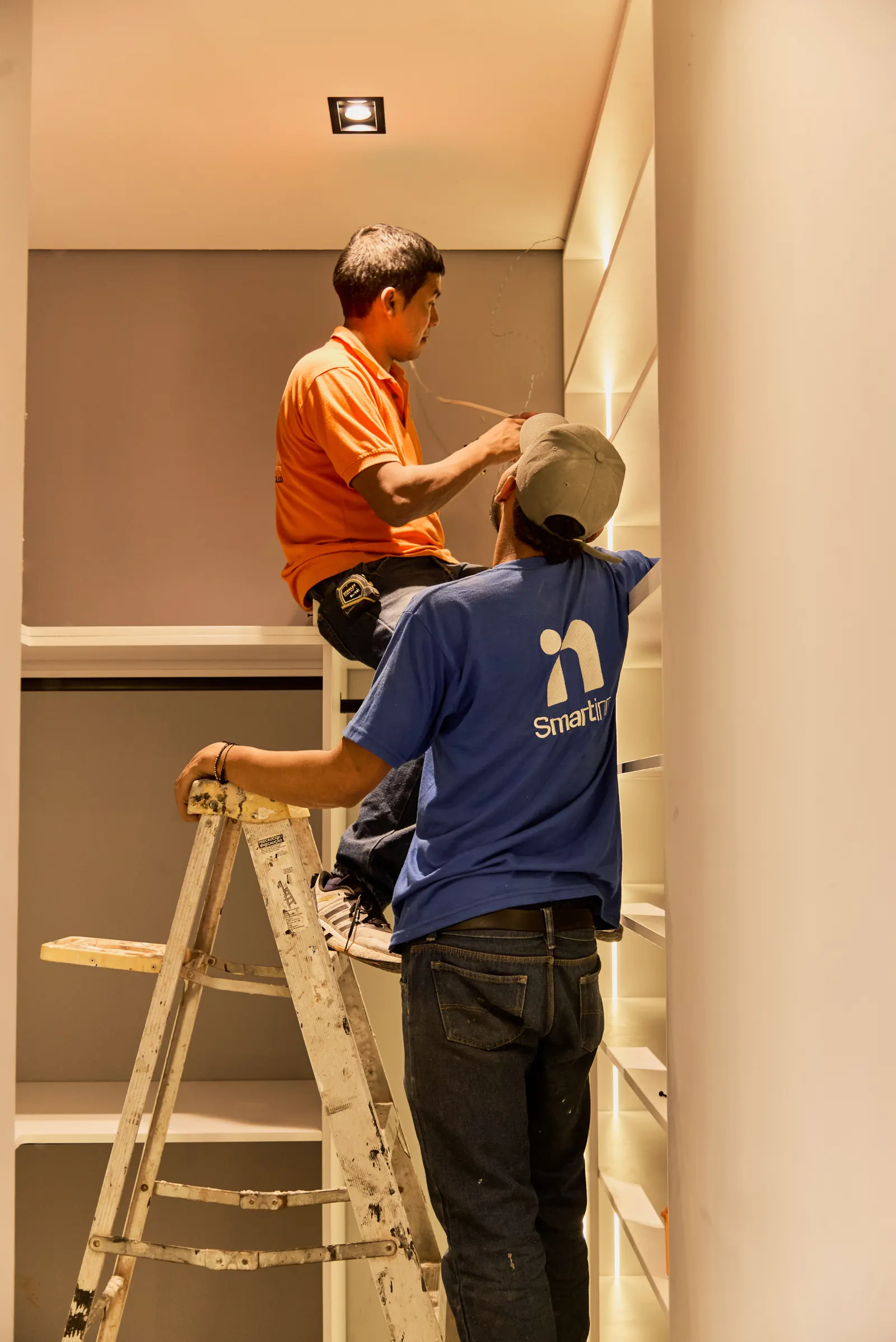 Dos carpinteros de Smartinn instalando los peldaños de una escalera de madera junto a una estantería iluminada