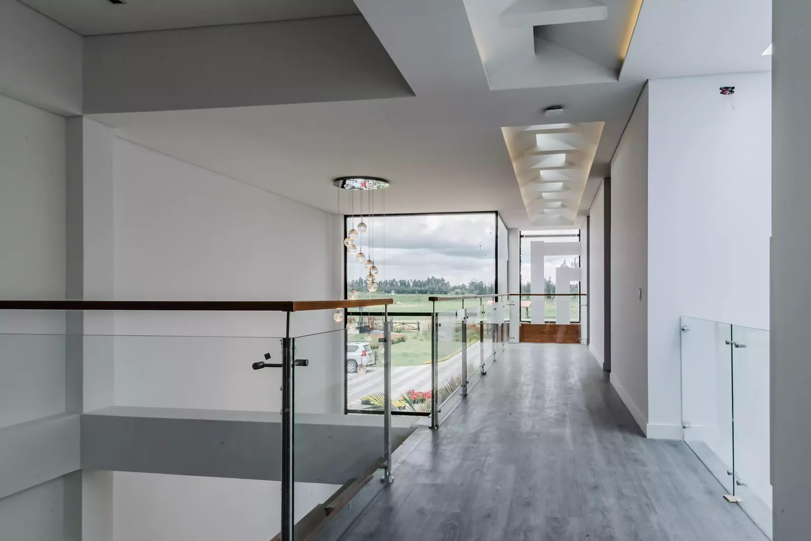Corredor del segundo piso de una casa, con baranda de vidrio, cielo raso con luz perimetral y un ventanal al fondo