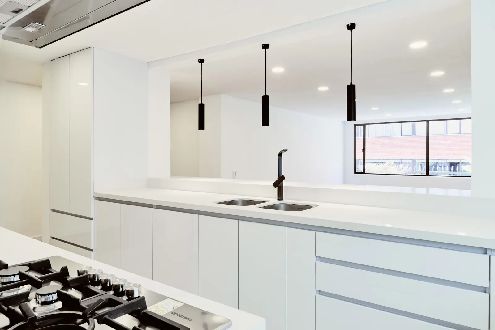 Cocina en L con muebles blancos a medida, mesón claro, lavaplatos doble y lámparas colgantes negras