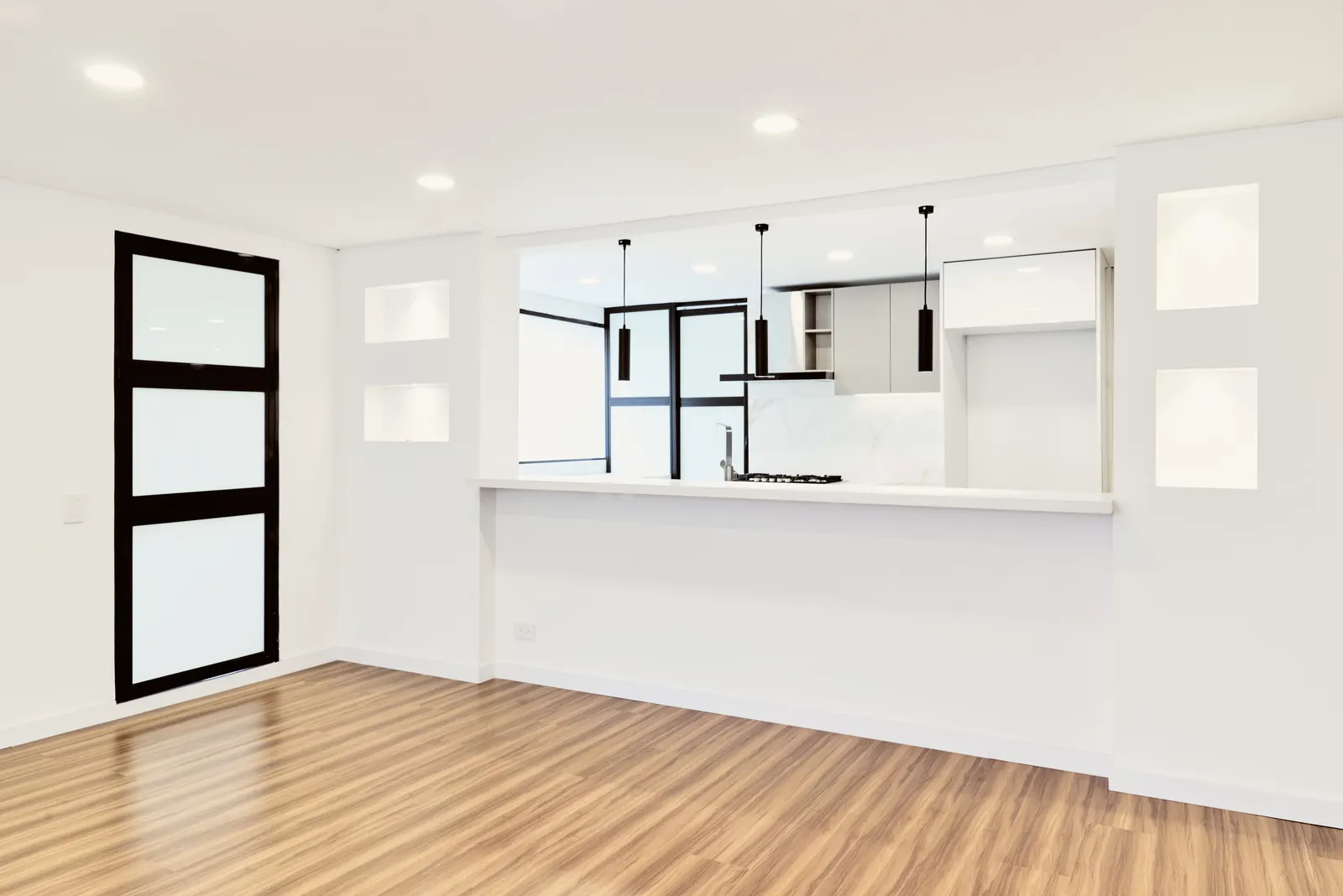 Sala con piso de madera, ventanal de marco negro y cocina abierta con barra blanca y lámparas colgantes