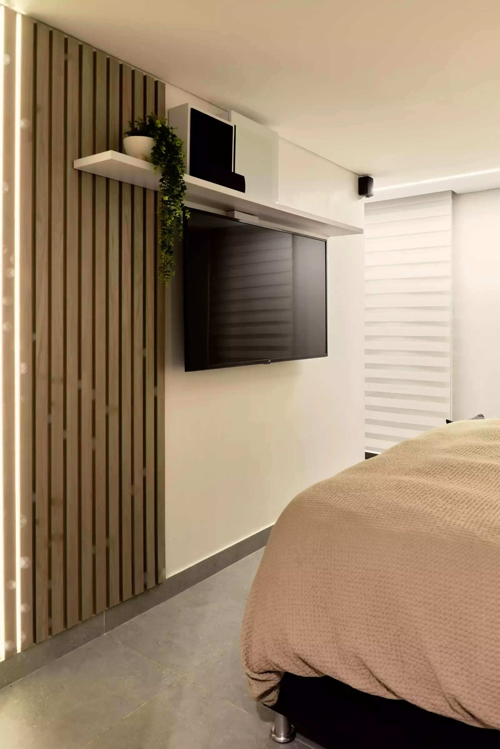 Televisión sobre un muro de listones de madera con luz lineal junto a la cama de un dormitorio