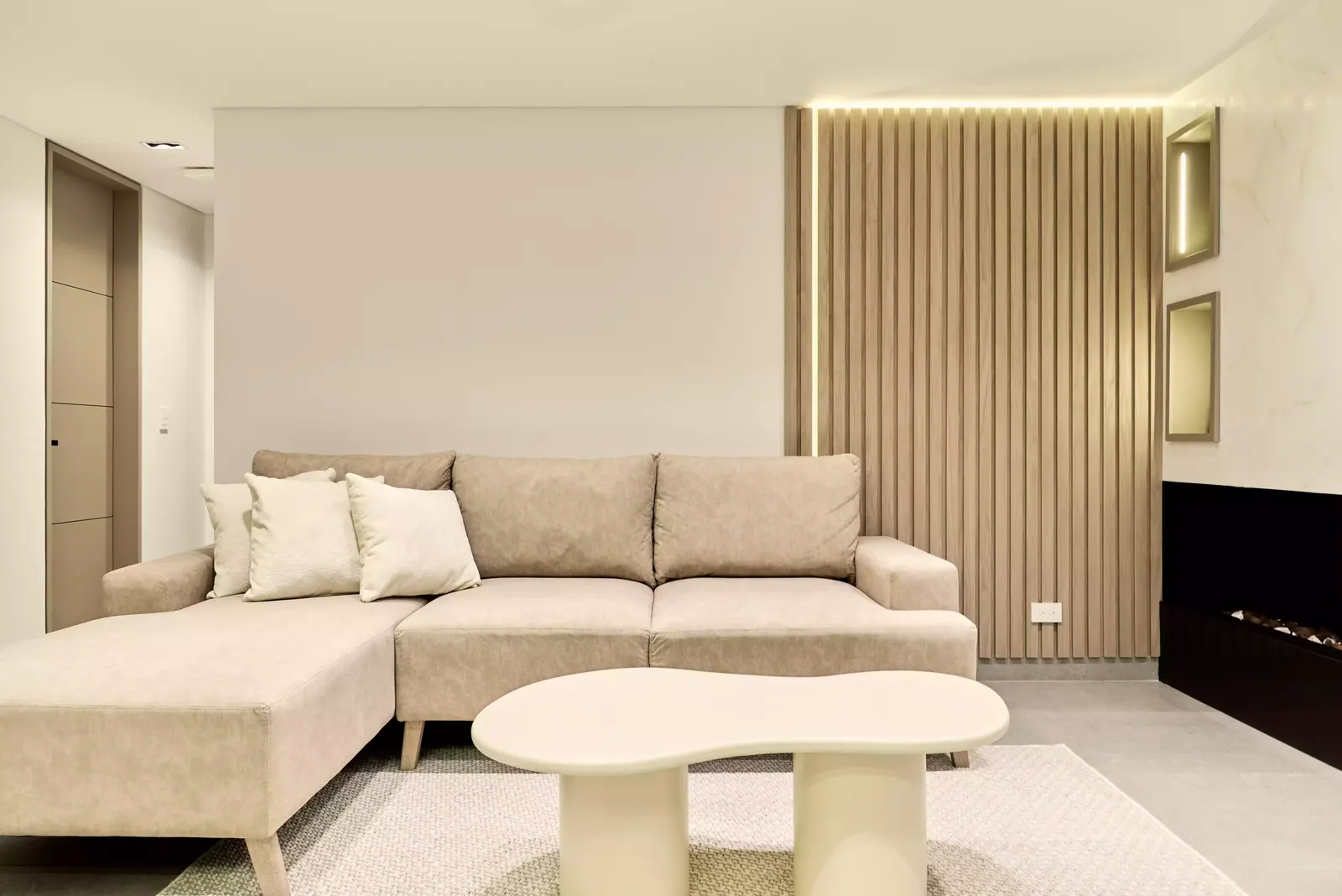 Sala de apartamento en tonos beige con muro de listones de madera, luz lineal y chimenea lineal