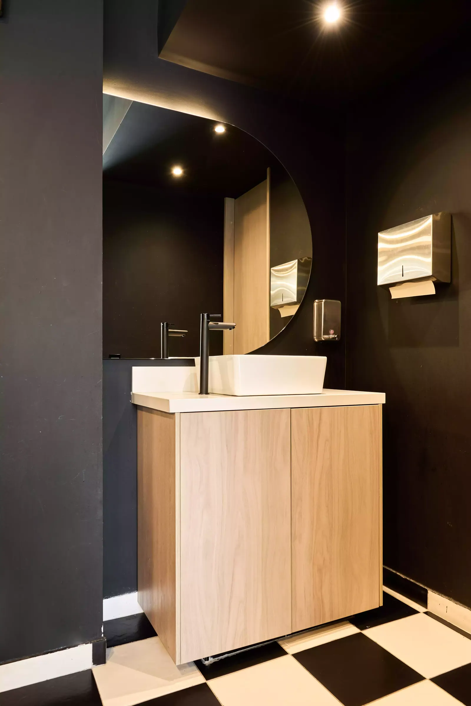 Baño con mueble de lavamanos de madera clara, espejo redondo y muro oscuro en un local comercial
