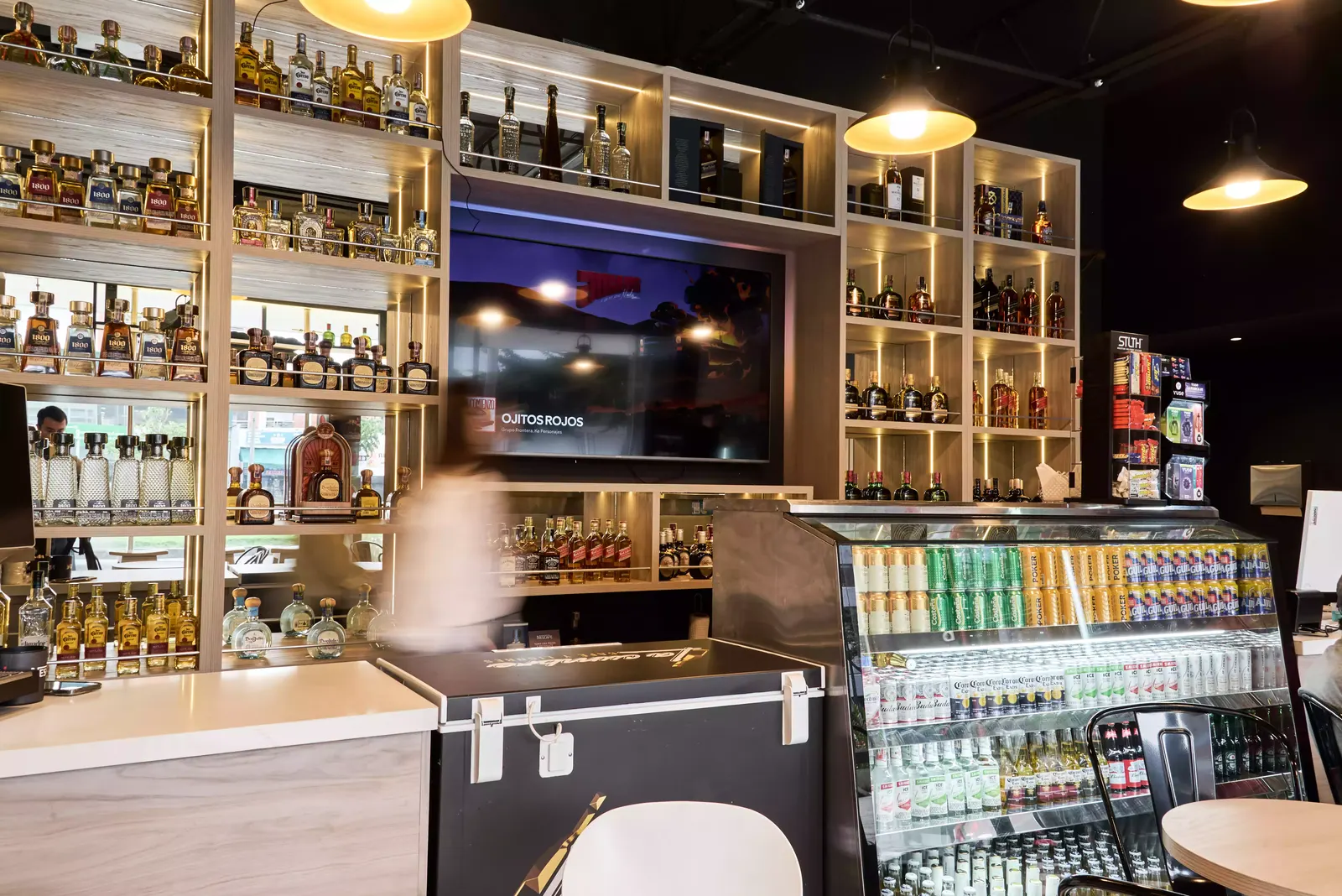 Estanterías de madera clara con botellas, pantalla integrada y luz en cada nivel, sobre la barra de un local comercial