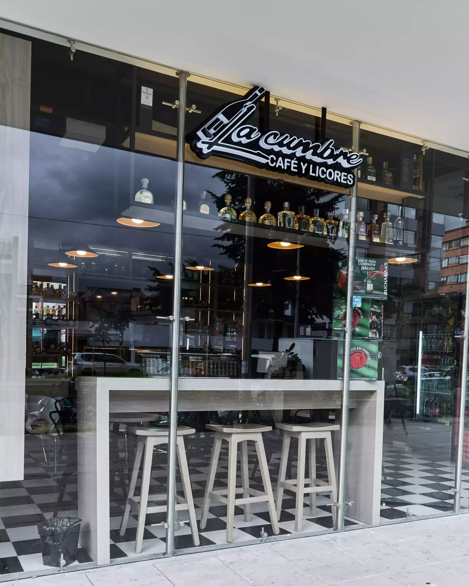 Fachada de vidrio de un local con el aviso La Cumbre café y licores y una barra con taburetes al frente