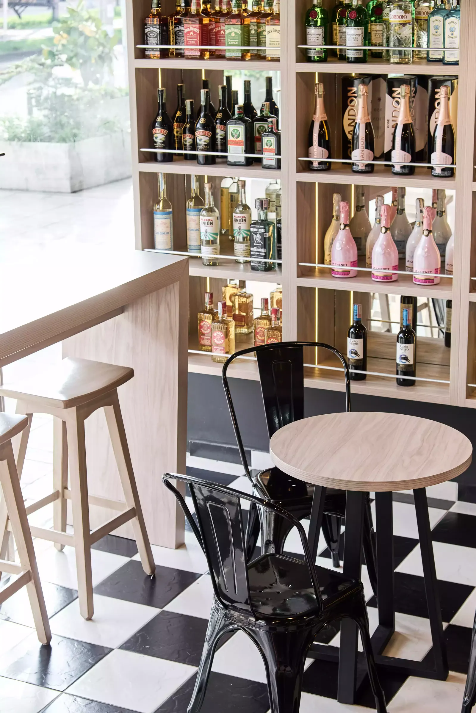 Mesa alta de madera clara con taburetes frente a las estanterías de licores de un local comercial