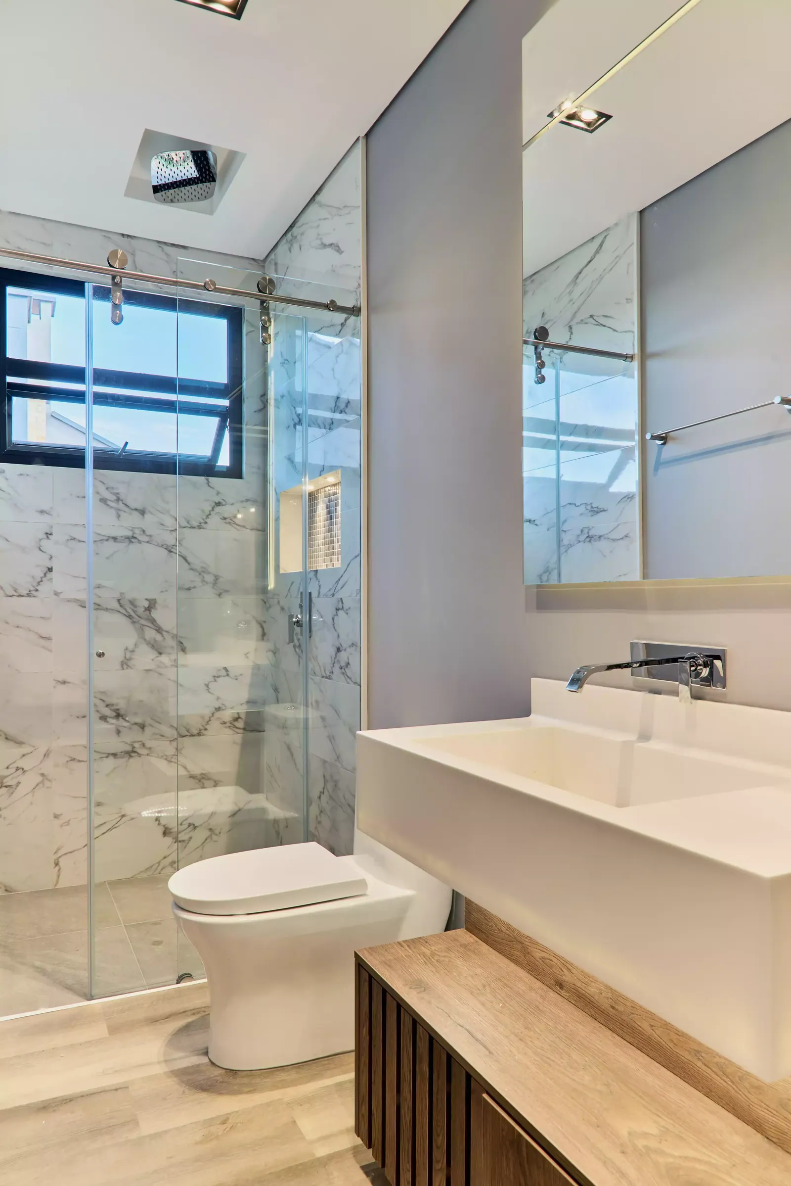 Baño con ducha en vidrio y revestimiento claro con veta, lavamanos rectangular sobre mueble suspendido y banco de madera