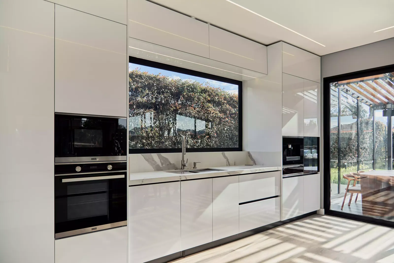 Cocina blanca de alto brillo con hornos empotrados y ventana corrida sobre el mesón