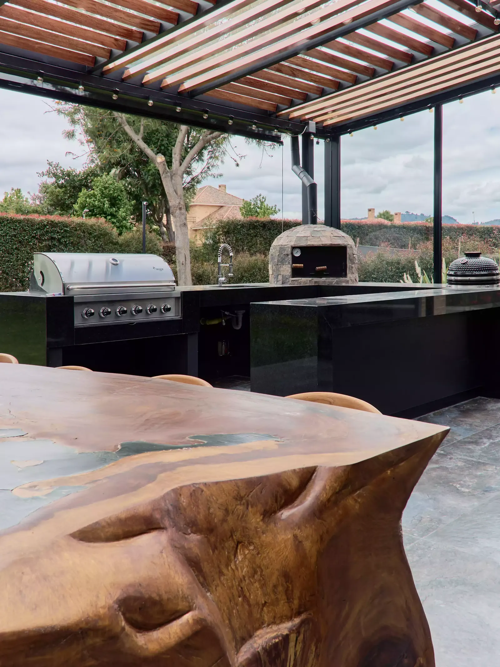 Cocina exterior bajo una pérgola de madera, con parrilla, horno de pizza y mesón negro junto a la mesa de madera