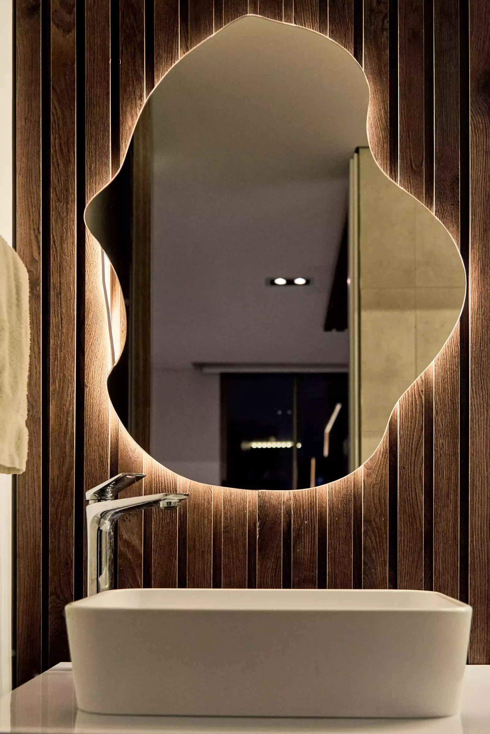 Baño con espejo de forma orgánica retroiluminado sobre un muro de listones de madera