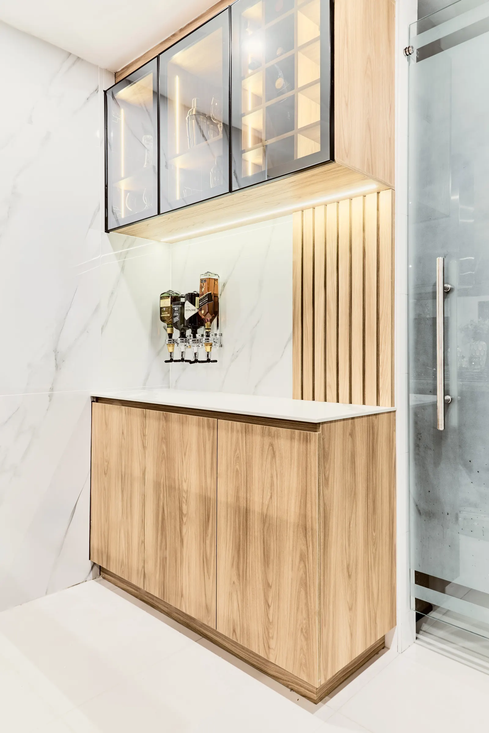 Mueble bar de madera clara con dispensadores de licor sobre un revestimiento claro con veta y vitrina superior