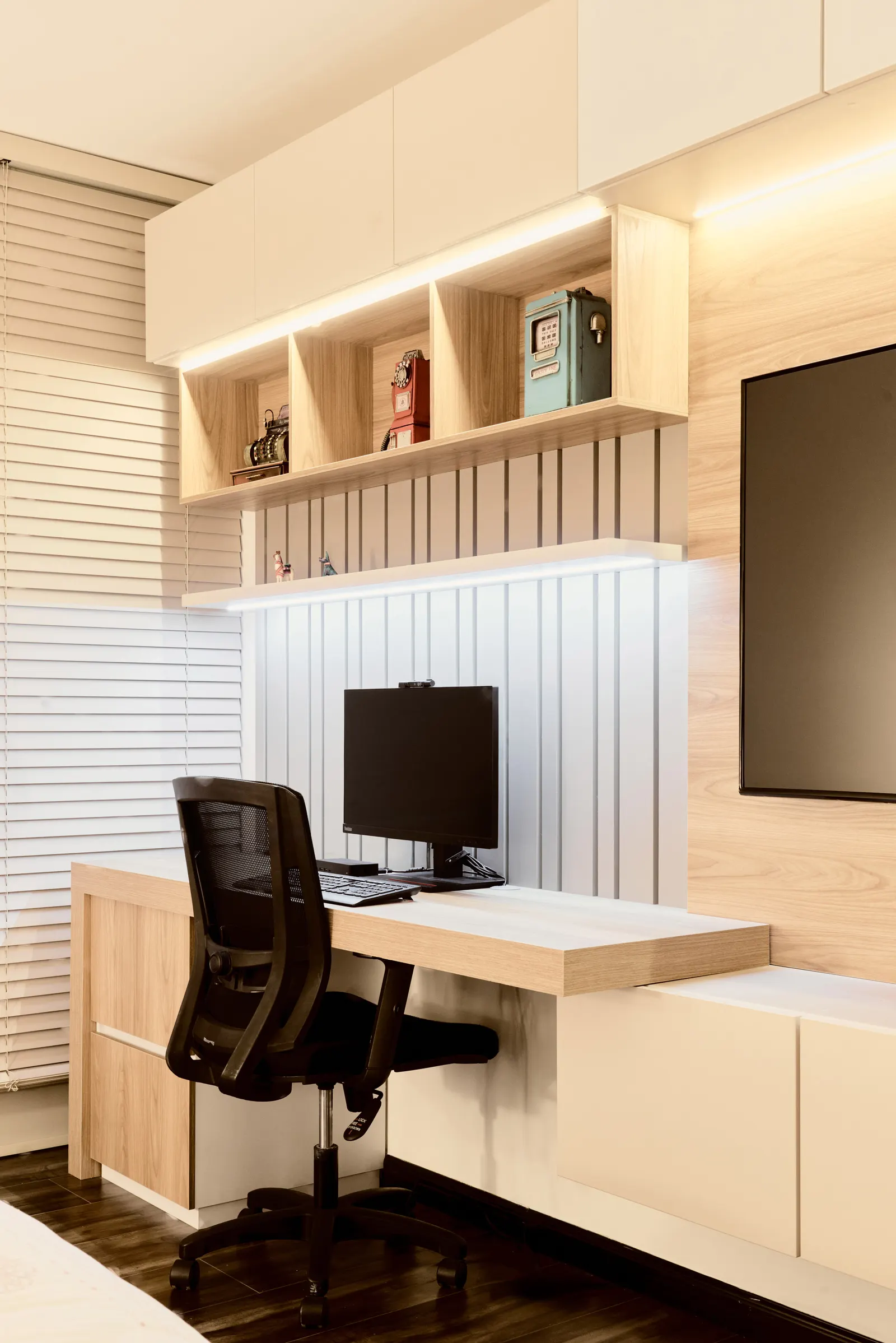Oficina en casa con escritorio, repisas de madera clara y luz lineal, junto a una pantalla